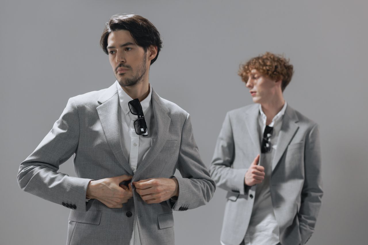 Studio shot of two men in gray suits posing confidently with sunglasses indoors.